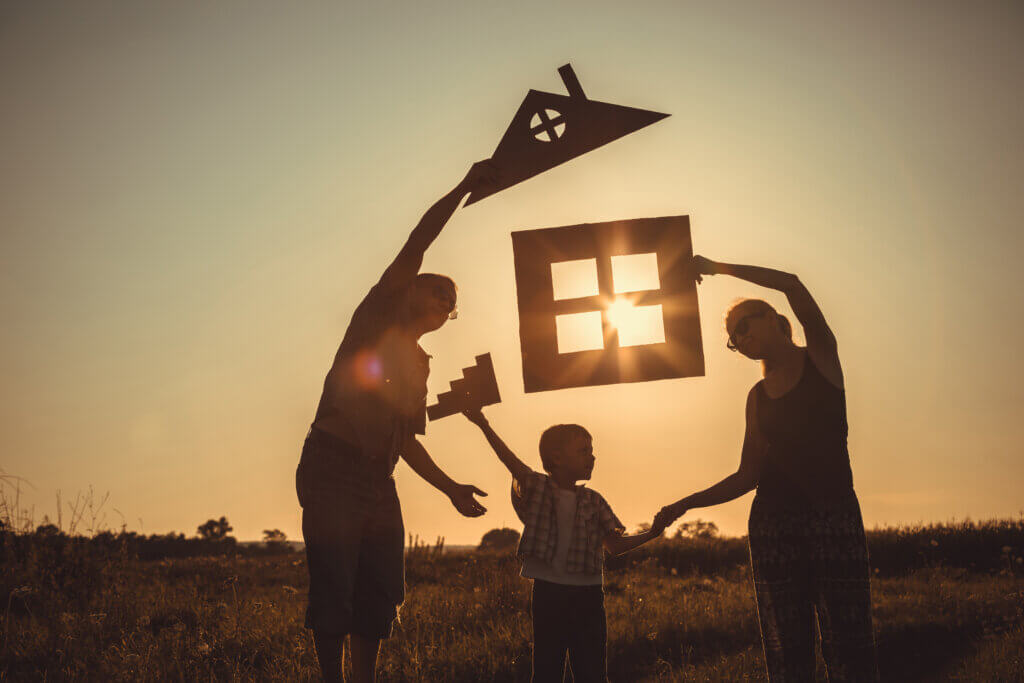 two adults and a child holding cutout of a house against a sunrise background